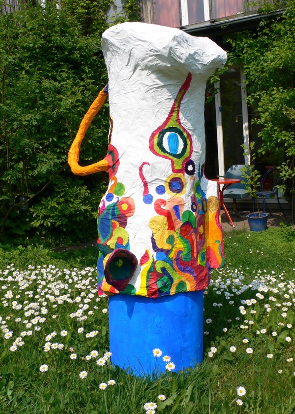 Bunte Gartenskulptur aus Pappmaché auf blauem Sockel in einer Blumenwiese