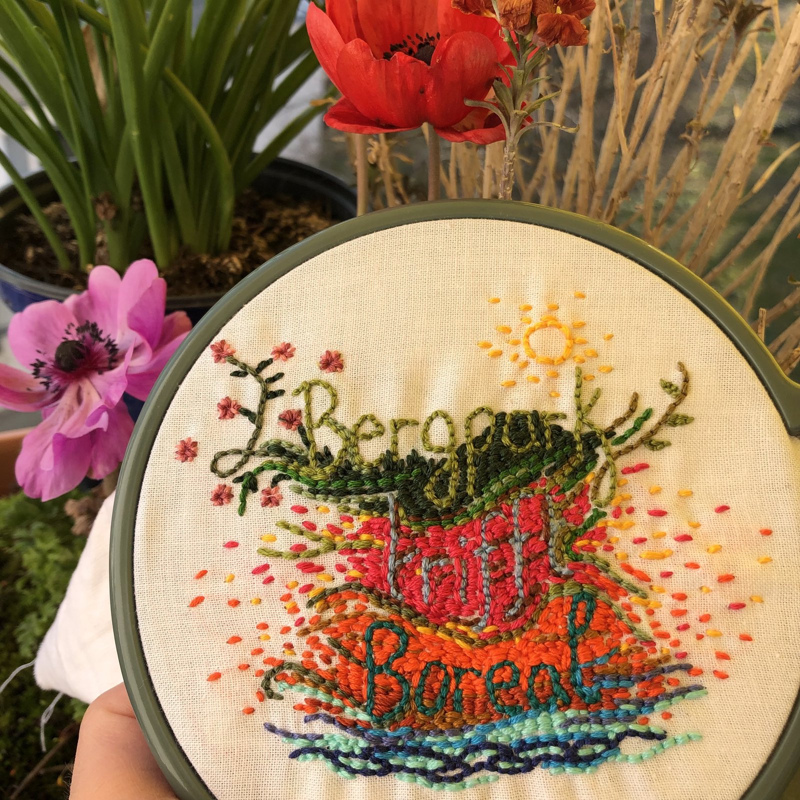 Stickrahmen mit bunter Stickerei eines Cafés, umgeben von Blumentöpfen